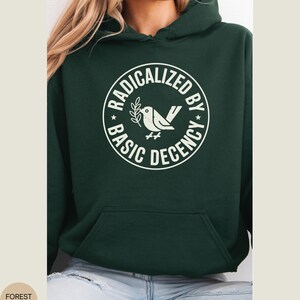 May include: Forest green hoodie with a white graphic. The graphic is a circle with the words "RADICALIZED BY BASIC DECENCY" around a dove holding a sprig. The hoodie has a front pocket and a hood.