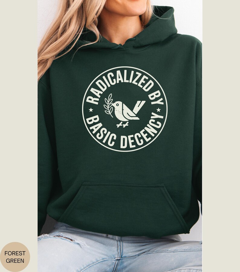 May include: Forest green hoodie with a white graphic. The graphic is a circle with the words "RADICALIZED BY BASIC DECENCY" around a dove holding a sprig. The hoodie has a front pocket and a hood.