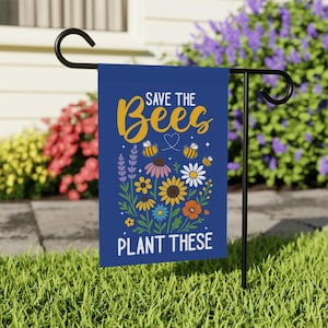 Può includere: Una bandiera da giardino blu con la scritta "SAVE THE Bees PLANT THESE" in oro e bianco. La bandiera presenta illustrazioni di api, fiori e un cuore. La bandiera è esposta su un supporto in metallo nero in un giardino.