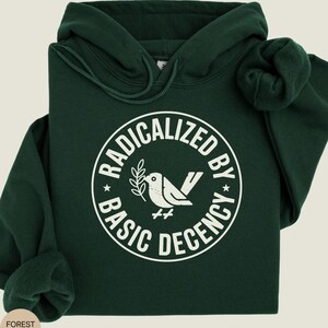May include: Forest green hoodie with a white graphic. The graphic is a circle with the words "RADICALIZED BY BASIC DECENCY" around a dove holding a sprig. The hoodie has a drawstring hood and a kangaroo pocket.