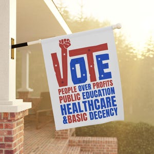 May include: A white rectangular garden flag featuring the word VOTE in large red and blue letters, with the letter V replaced by a raised fist. Below, the text reads PEOPLE OVER PROFITS, PUBLIC EDUCATION, HEALTHCARE, & BASIC DECENCY in red and blue.
