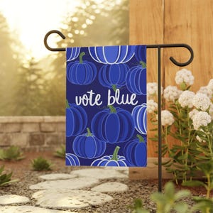 May include: A rectangular garden flag featuring a dark blue background with a repeating pattern of various blue pumpkins in different shades. The text "vote blue" is printed in white, lowercase, cursive script across the center of the flag.