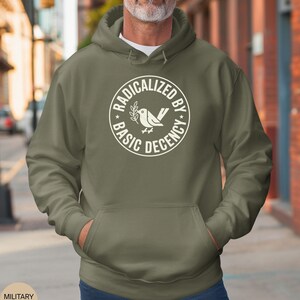 May include: An olive green hoodie with a front pocket and a drawstring hood. The white graphic on the chest reads "RADICALIZED BY BASIC DECENCY" in a circular design with a bird and olive branch illustration.