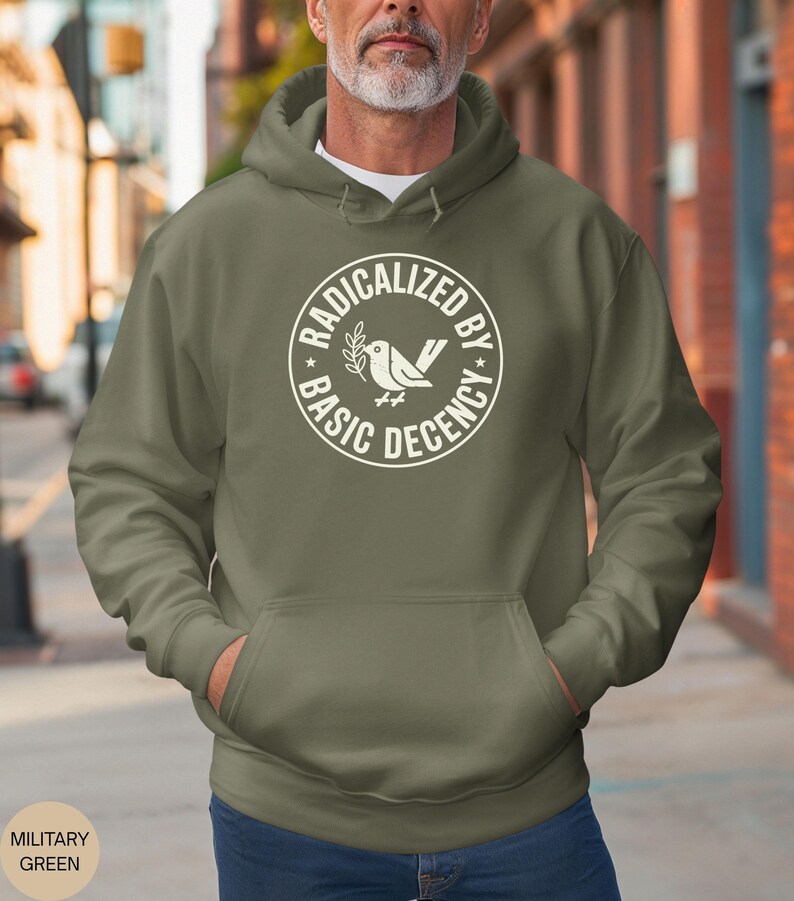 May include: An olive green hoodie with a front pocket and a drawstring hood. The white graphic on the chest reads "RADICALIZED BY BASIC DECENCY" in a circular design with a bird and olive branch illustration.