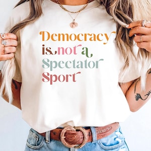 Può includere: T-shirt color crema con la scritta "Democracy is not a Spectator Sport" in lettere arancioni, rosa, verdi e rosse. La maglietta è abbinata a jeans blu e una cintura marrone con fibbia decorativa.