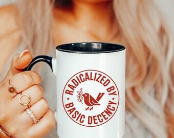 May include: White ceramic mug with a black handle and rim. The mug features a red circular design with the words "RADICALIZED BY BASIC DECENCY" surrounding a bird illustration holding an olive branch. The mug is held by a person.
