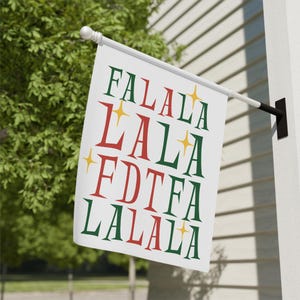 Peut inclure: Un drapeau blanc avec les mots "FALALA" en vert et rouge, avec des étoiles jaunes. Le drapeau est monté sur un poteau blanc et est fixé à un mur blanc. Le drapeau est affiché à l'extérieur, avec un feuillage vert en arrière-plan.