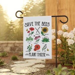 Può includere: Una bandiera da giardino bianca con la scritta "SAVE THE BEES" e "=PLANT THESE=". La bandiera presenta illustrazioni di varie piante come papaveri, lavanda e girasoli, con i nomi di ogni pianta sottostanti. La bandiera è esposta all'aperto.
