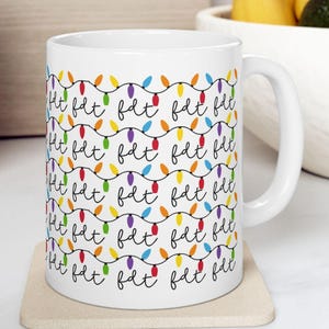 May include: White ceramic mug with a repeating pattern of colorful Christmas lights and the word "fat" in a handwritten font. The mug sits on a beige coaster.