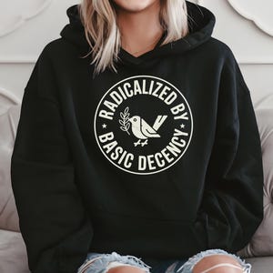 May include: Black hooded sweatshirt with a white circular graphic. The graphic features a bird with a sprig and the words "RADICALIZED BY BASIC DECENCY" in a circular design. The sweatshirt is worn by a person with blonde hair and blue jeans.