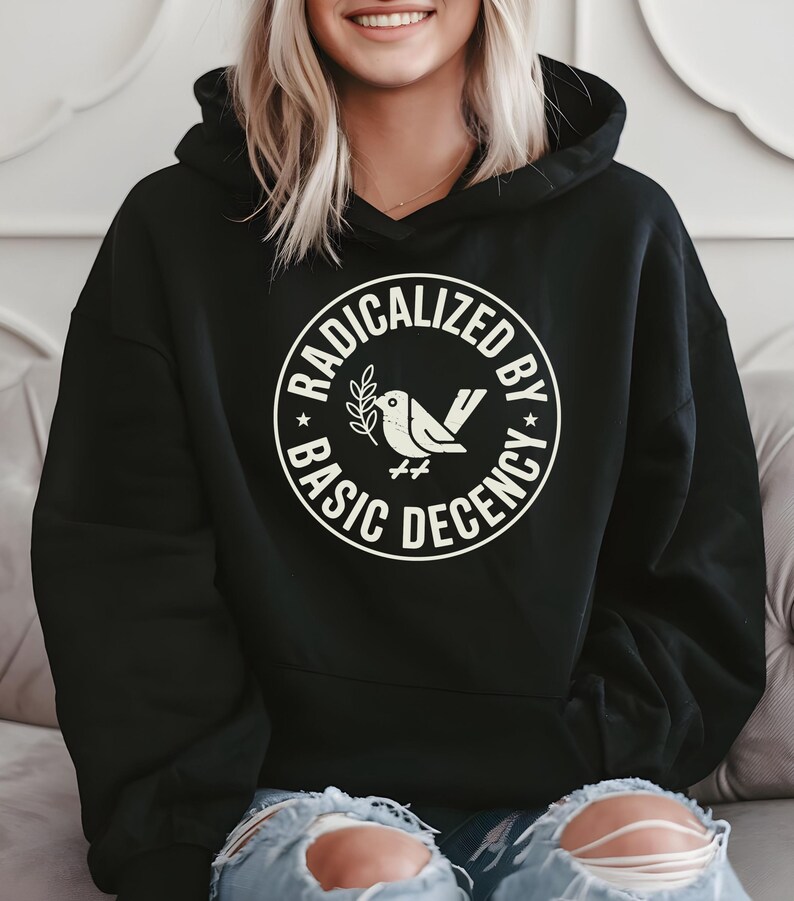 May include: Black hooded sweatshirt with a white circular graphic. The graphic features a bird with a sprig and the words "RADICALIZED BY BASIC DECENCY" in a circular design. The sweatshirt is worn by a person with blonde hair and blue jeans.