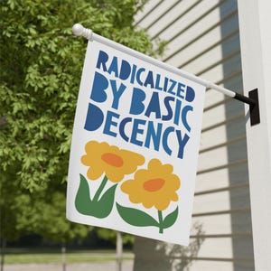 Op de afbeelding: Een witte vlag met de tekst "RADICALIZED BY BASIC DECENCY" in blauw. Twee gele bloemen met oranje centra en groene bladeren staan onder de tekst. De vlag is gemonteerd op een witte muur naast groen gebladerte.