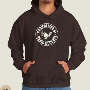 May include: Dark chocolate-colored hoodie with a white circular graphic that reads "RADICALIZED BY BASIC DECENCY" around a bird and olive branch design. The hoodie has a hood and a front pocket.