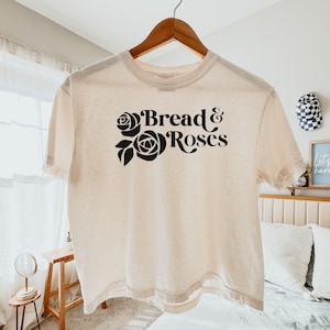 Puede incluir: Camiseta color crema con el texto "Bread & Roses" en negro, con detalles florales. La camiseta está colgada en una percha de madera. Es una prenda de vestir informal.