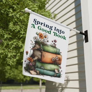 May include: A white flag with the text "Spring Into A Good Book" in navy and green. The flag features a stack of books with flowers and greenery growing from them. The flag is hanging outside.