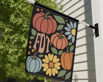 May include: Black outdoor flag featuring a fall-themed design with pumpkins, flowers, and the letters "FDT" in a retro font. The pumpkins are in shades of orange and the flowers are blue and yellow. The flag is hanging on a white wall.