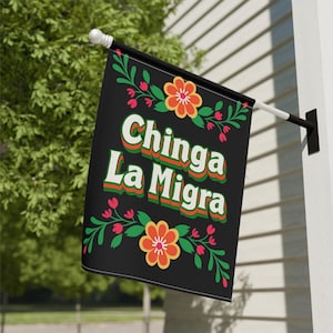 May include: Black flag with the text "Chinga La Migra" in white, green, and orange lettering. The flag is decorated with floral designs in red, green, and orange. The flag is hanging outside on a white wall.