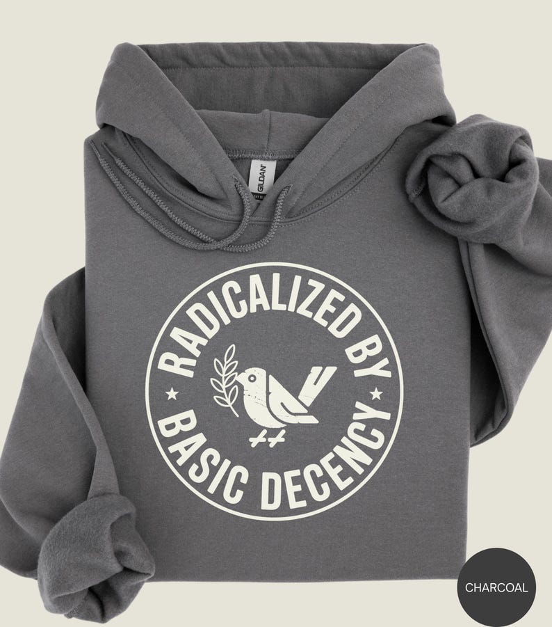 May include: Charcoal gray hoodie with a white circular graphic. The graphic reads "RADICALIZED BY BASIC DECENCY" around a bird holding an olive branch. The hoodie has a drawstring hood and a kangaroo pocket.