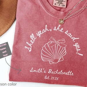 May include: Crimson t-shirt with the text "Shell yeah, she said yes!" and "Smith's Bachelorette Est. 2025". The shirt features a seashell graphic and a Comfort Colors tag. A gold necklace is draped across the shirt.