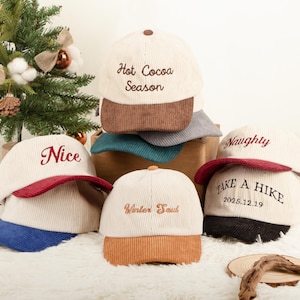 Könnte beinhalten: Eine Auswahl an cremefarbenen Cord-Baseballkappen mit kontrastierenden Schirmen und gestickten Texten. Die Kappen zeigen Sätze wie "Hot Cocoa Season", "Nice" und "Take a Hike 2025.12.19". Winterliches Thema.