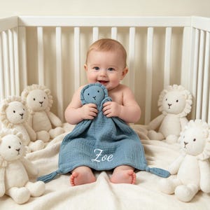 Könnte beinhalten: Ein lächelndes Baby hält eine blaue Schmusedecke mit dem weißen Namen "Zoe" in einem Kinderbett. Mehrere weiße Löwen-Stofftiere umgeben das Baby auf einer cremefarbenen Decke.
