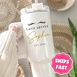 May include: White tumbler with a handle, featuring the text "LASH ARTIST Sophia EST. 2024" in black and gold script. The design includes eyelash graphics. The tumbler has a clear lid and a pink "SHIPS FAST" sticker.