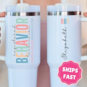 May include: Two white insulated tumblers with handles. One features the text "registered BEHAVIOR technician" in colorful letters. The other has "RB" and the name "Elizabeth". Both have a lid and a straw.