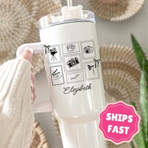 May include: White insulated tumbler featuring black postage stamp designs of cameras, film reels, and a clapperboard. The name "Elizabeth" is written in cursive. The tumbler has a white handle and a clear lid.