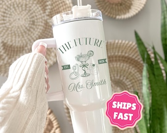 May include: White tumbler with a handle, featuring the text "THE FUTURE" and "Mrs. Smith" in green script. The design includes illustrations of a cocktail, rings, and a drink. The tumbler has a clear lid and a straw.