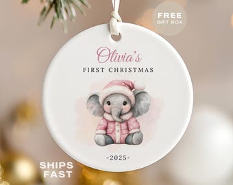May include: A white ceramic Christmas ornament with a watercolor illustration of a baby elephant wearing a pink coat and hat. The ornament reads "Olivia's First Christmas - 2025".
