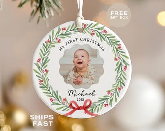 May include: A round white ceramic Christmas ornament featuring a scalloped center window displaying a photograph of a smiling baby. The ornament is decorated with a green holly wreath, red berries, a red bow, and the text My First Christmas, Michael, 2025.