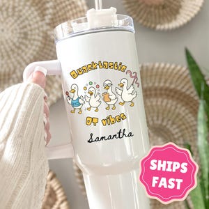 May include: White tumbler with a handle, featuring a cartoon duck design with the text "Quacktastic OT vibes Samantha." The design includes ducks wearing vests and holding objects. A pink sticker that says "SHIPS FAST" is visible.