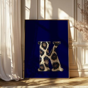 May include: A framed print displaying the letter 'K' in a leopard print pattern, set against a dark blue backdrop. The artwork is enclosed within a gold frame. The print is displayed in a room, suggesting a decorative home accent. The frame's dimensions are in centimetres.