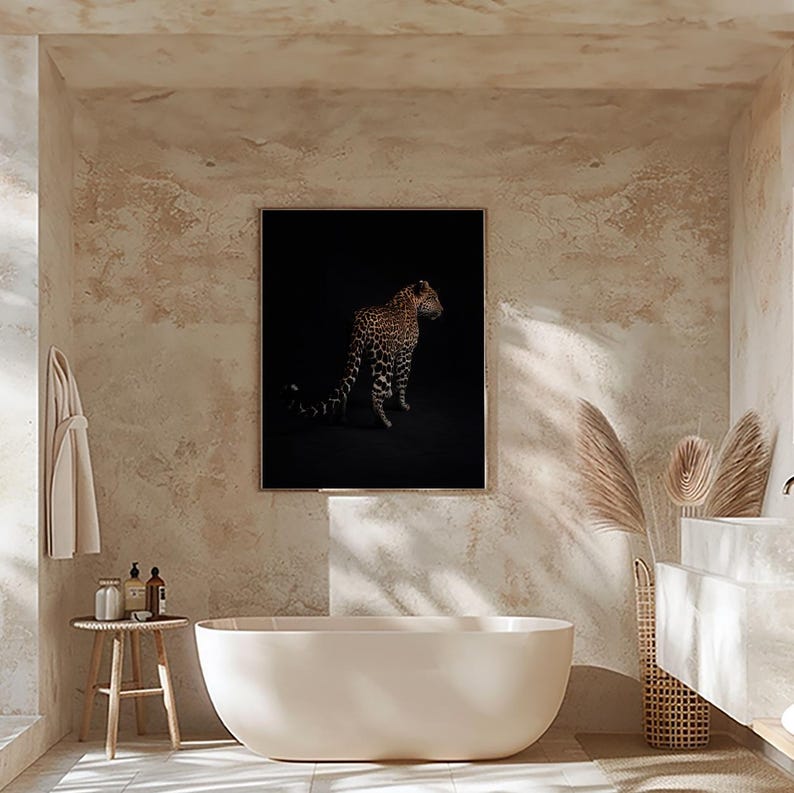 May include: A black and white photograph of a leopard standing in a dark setting. The leopard is facing to the right and has a spotted coat. The photograph is framed and hanging on a beige wall in a bathroom.