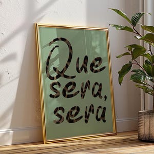 Peut inclure: Impression d'art encadrée avec l'inscription "Que sera, sera" dans une police élégante. Le texte présente un motif léopard sur un fond vert clair. Le cadre est doré, ajoutant une touche d'élégance à l'œuvre.