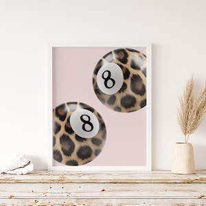May include: A framed art print showing two pool balls with a leopard print pattern. Each ball has a white circle with the number 8 in black. The background is a pale pink colour.