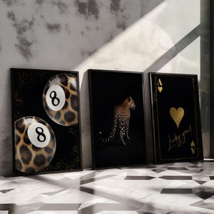 May include: Three framed prints with black backgrounds and gold accents. The left print features two 8-ball pool balls with a leopard print design. The middle print features a leopard in a black and white silhouette. The right print features a gold ace of hearts playing card with the text "Lucky you!" in gold script.