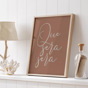 May include: A framed print with a terracotta background and white cursive text that reads "Que sera sera".
