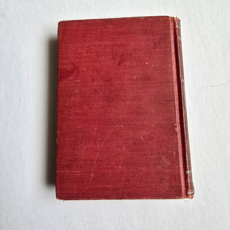 Peut inclure: Un livre ancien &agrave; couverture rigide rouge d&eacute;lav&eacute;. La tranche du livre est visible, avec le titre et l'auteur. La couverture pr&eacute;sente un aspect textur&eacute; avec une l&eacute;g&egrave;re usure, sugg&eacute;rant l'&acirc;ge et l'utilisation. Le livre est pos&eacute; sur un fond blanc.