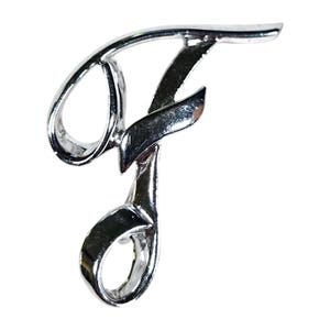 May include: A shiny, silver-colored metal letter "F" in a cursive script. The letter is highly polished, reflecting light and creating highlights. The design is a single, continuous line, with a loop at the top and bottom.