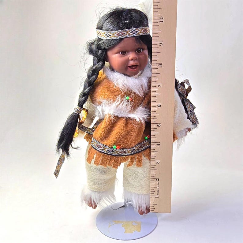 May include: A Native American doll, approximately 10 inches tall, with black hair in a braid and wearing a traditional outfit. The doll's outfit includes a brown tunic with white faux fur trim, beaded accents, and matching pants. A headband with geometric patterns adorns the doll's head.