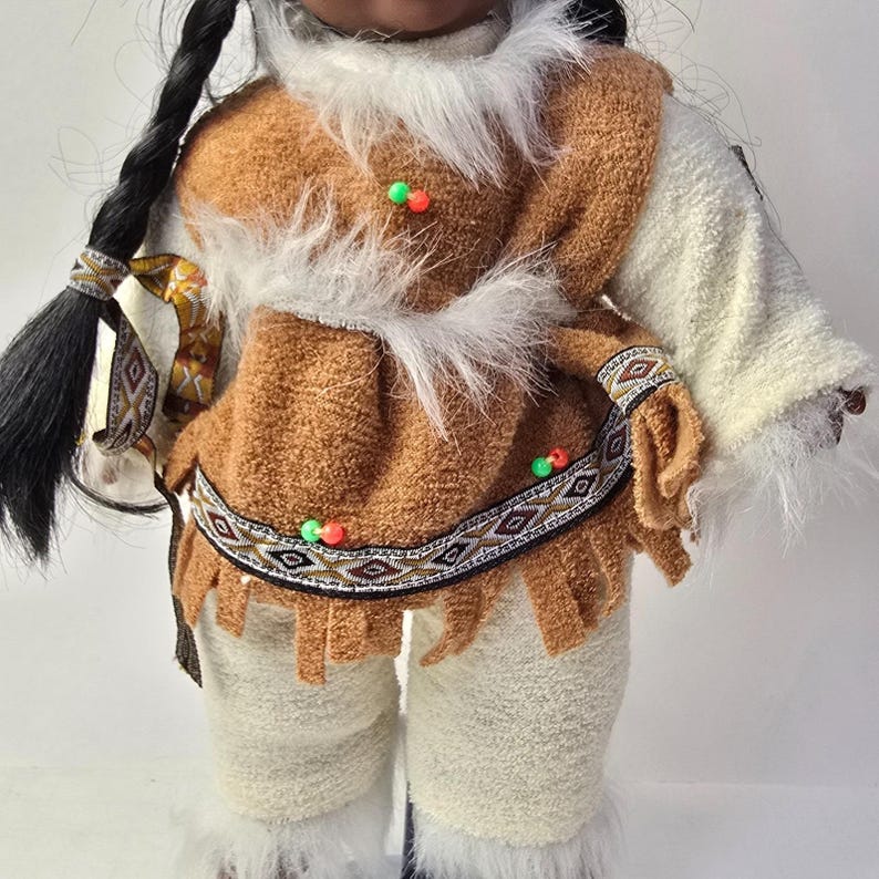 May include: A doll dressed in traditional Native American attire. The doll wears a brown vest with white faux fur trim, a patterned belt, and white pants with faux fur trim. The doll has long black braids and is adorned with colorful beads.