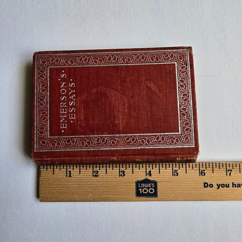 Peut inclure: Un petit livre rectangulaire avec une couverture rouge et une bordure d&eacute;corative argent&eacute;e. Le texte "EMERSON'S ESSAYS" est imprim&eacute; en blanc. Une r&egrave;gle en bois est plac&eacute;e &agrave; c&ocirc;t&eacute; du livre.