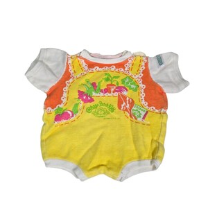 May include: A vintage Cabbage Patch Kids baby romper. The romper is yellow with an orange bib featuring a garden scene and the Cabbage Patch Kids logo. White sleeves and trim complete the design.