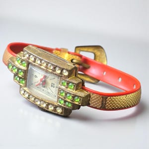 May include: Vintage rectangular wristwatch with a gold-tone case, adorned with clear and green rhinestones. The watch has a red leather strap with a gold buckle. The watch face is white with black numerals and red hands.