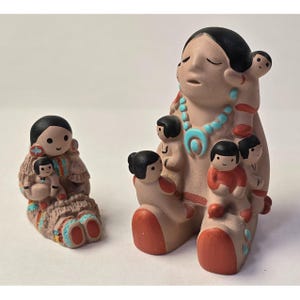 Puede incluir: Dos figuras de cerámica que representan una figura materna con varios niños. La figura más grande tiene un collar turquesa y detalles rojos. La figura más pequeña está sentada, también con niños. Ambas tienen cabello negro y están pintadas en tonos tierra.
