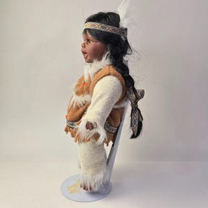 May include: A Native American doll with dark hair in a braid, wearing a brown and white outfit with fringe. The doll has a headband with a geometric pattern and a white feather. The doll is standing on a light blue stand.