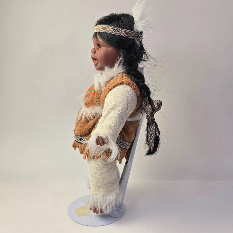 May include: A Native American doll with dark hair in a braid, wearing a brown and white outfit with fringe. The doll has a headband with a geometric pattern and a white feather. The doll is standing on a light blue stand.