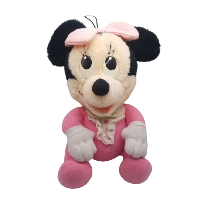 Puede incluir: Un juguete de peluche rosa de una Minnie Mouse beb&eacute; que lleva un vestido rosa con ribete de encaje blanco y un lazo rosa. El juguete tiene orejas negras y una nariz negra.