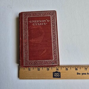 Peut inclure: Un petit livre ancien intitul&eacute; "Emerson's Essays" avec une couverture rouge et une bordure argent&eacute;e. Le livre est plac&eacute; &agrave; c&ocirc;t&eacute; d'une r&egrave;gle en bois, indiquant sa taille. La couverture du livre est l&eacute;g&egrave;rement us&eacute;e, ce qui sugg&egrave;re son &acirc;ge.
